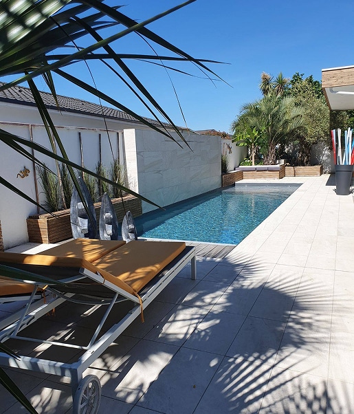 Piscine moderne rectangulaire avec transats orange, mur texturé blanc, et végétation luxuriante sous un ciel bleu ensoleillé.