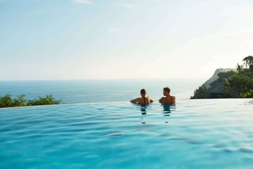 Dos d'un couple dans une piscine à débordement turquoise, admirant l'océan lointain et un paysage tropical vallonné avec des bungalows.