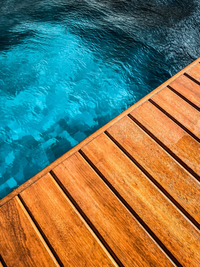 Piscine bleue éclatante et terrasse en bois exotique sous le soleil, séparées par une ligne diagonale nette. Atmosphère estivale.