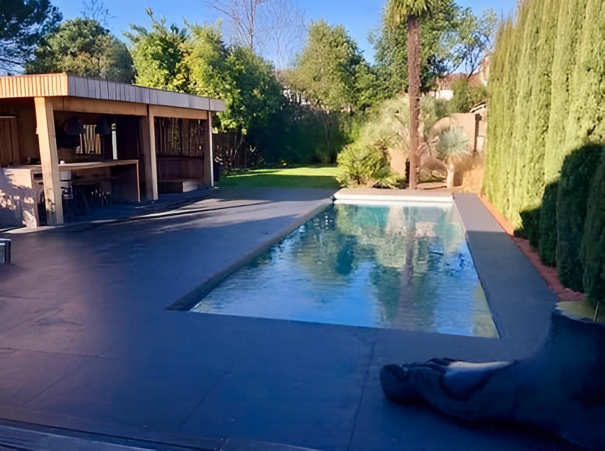 Piscine rectangulaire moderne avec terrasse foncée, pool house en bois et jardin verdoyant ensoleillé.