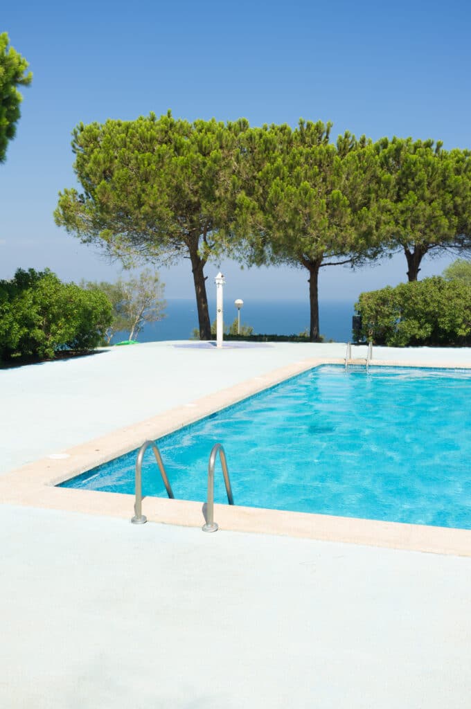 Piscine bleue ensoleillée avec échelles, bordée de pins. Vue panoramique sur la mer Méditerranée et ciel azur, idéal pour la détente.
